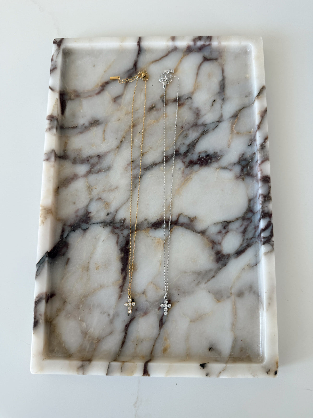 Two delicate cross pendant necklaces, one gold and one silver, displayed on a white marble jewelry tray with dark veining.