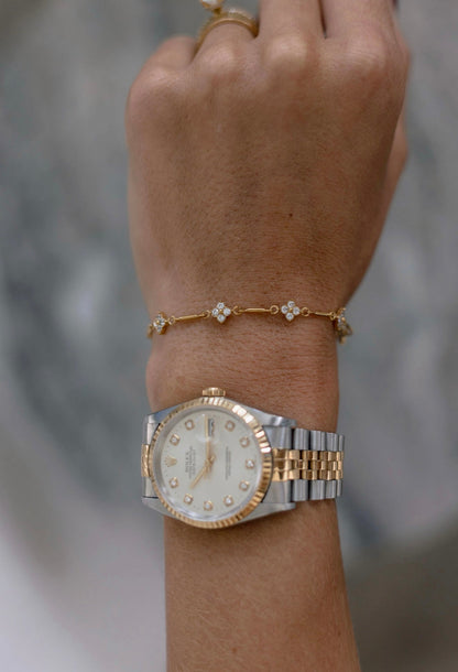 A close-up of a person's wrist wearing a two-tone Rolex watch with a white dial and diamond hour markers, along with a delicate gold bracelet, against a blurred gray background.