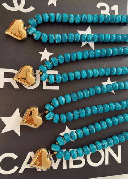 Four turquoise beaded necklaces with gold heart charms, arranged diagonally on a dark background with white star and text patterns.