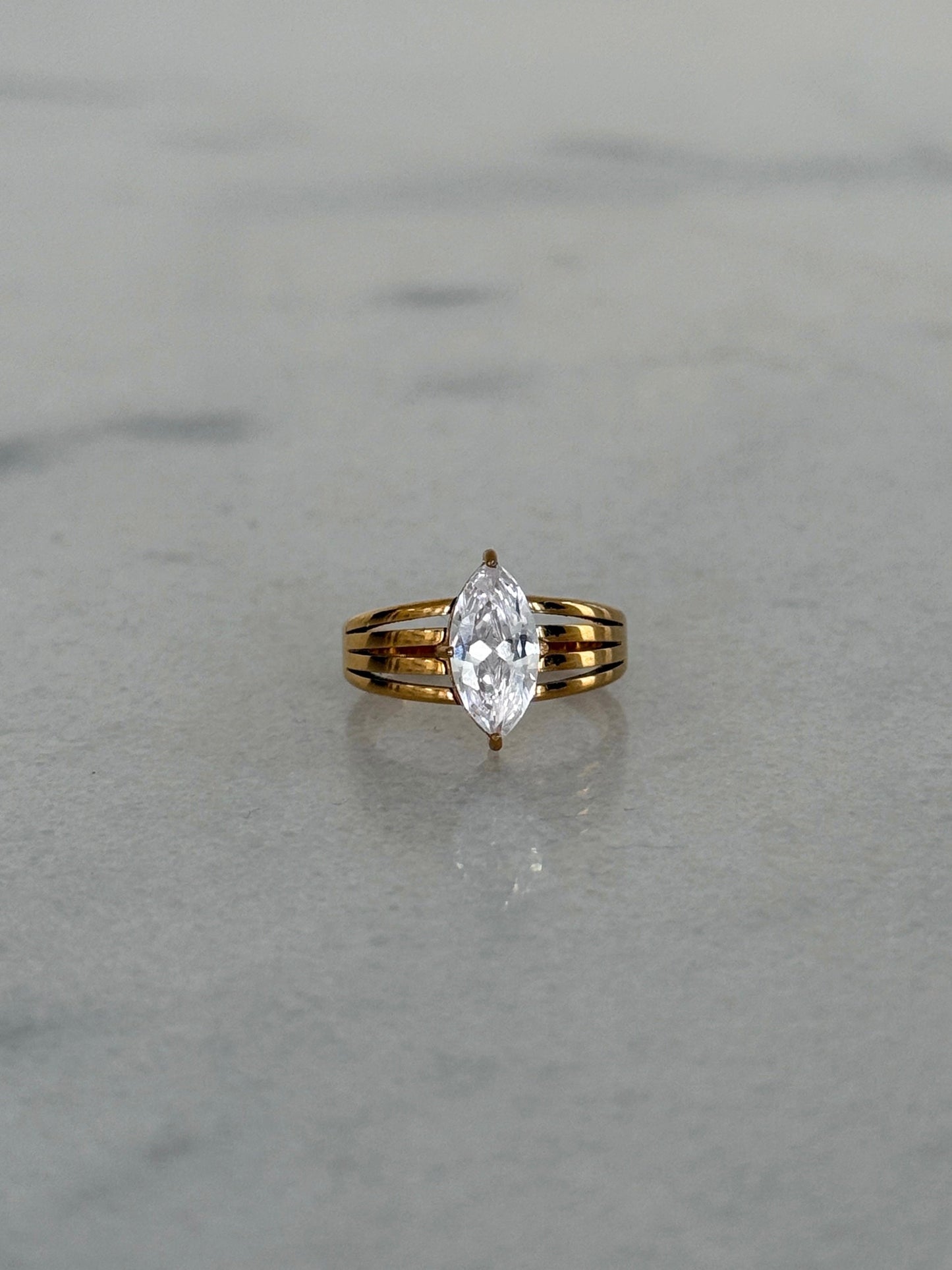 A yellow gold ring with a marquise-cut clear gemstone, set on a light gray marble surface.