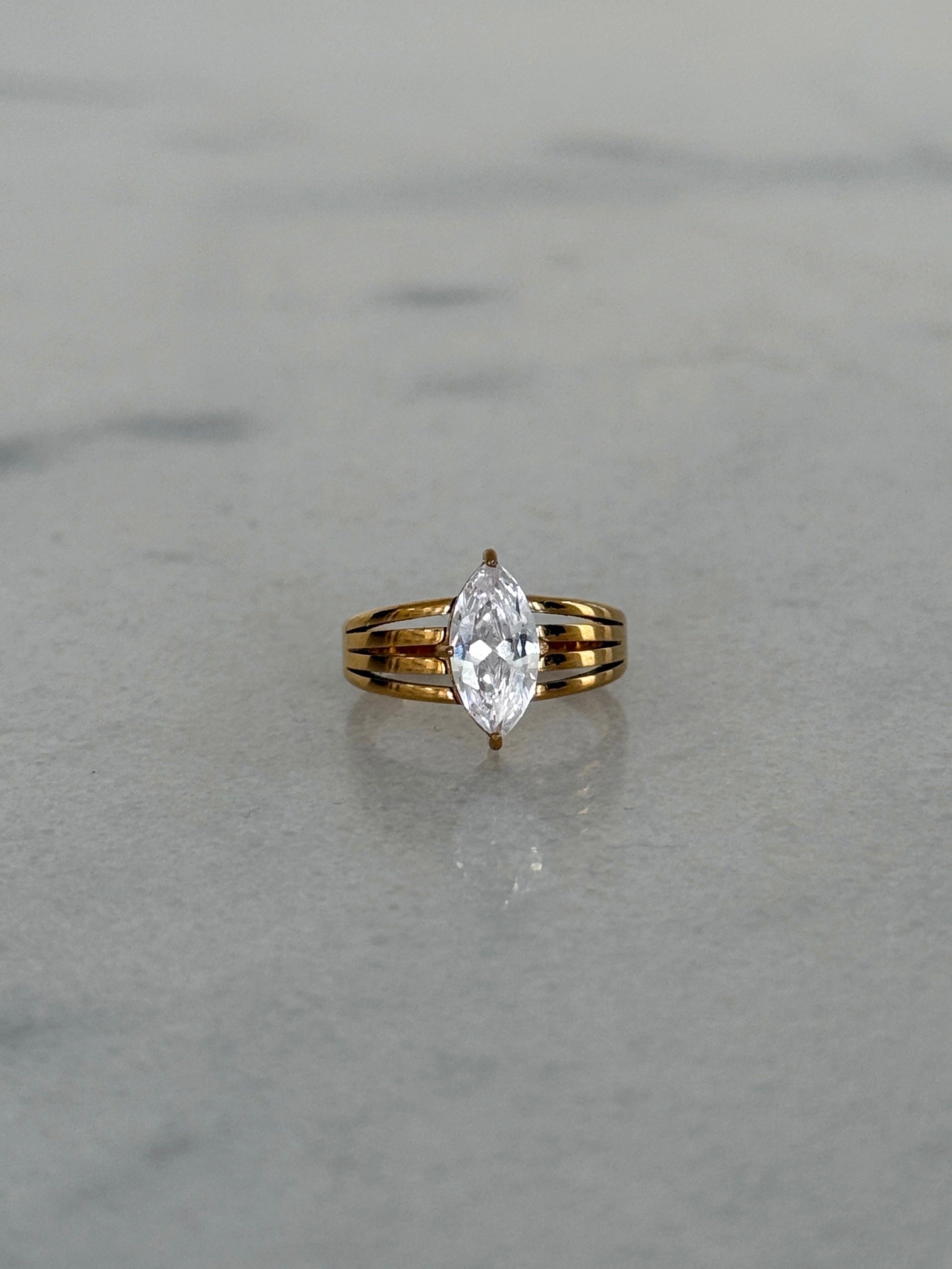 A yellow gold ring with a marquise-cut clear gemstone, set on a light gray marble surface.