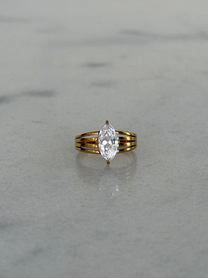 A yellow gold ring with a marquise-cut clear gemstone, set on a light gray marble surface.