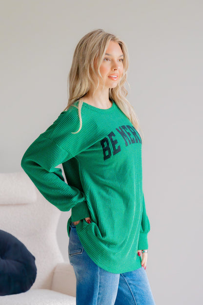 Woman wearing a green sweater with 'BE MINE' text, standing against a plain background