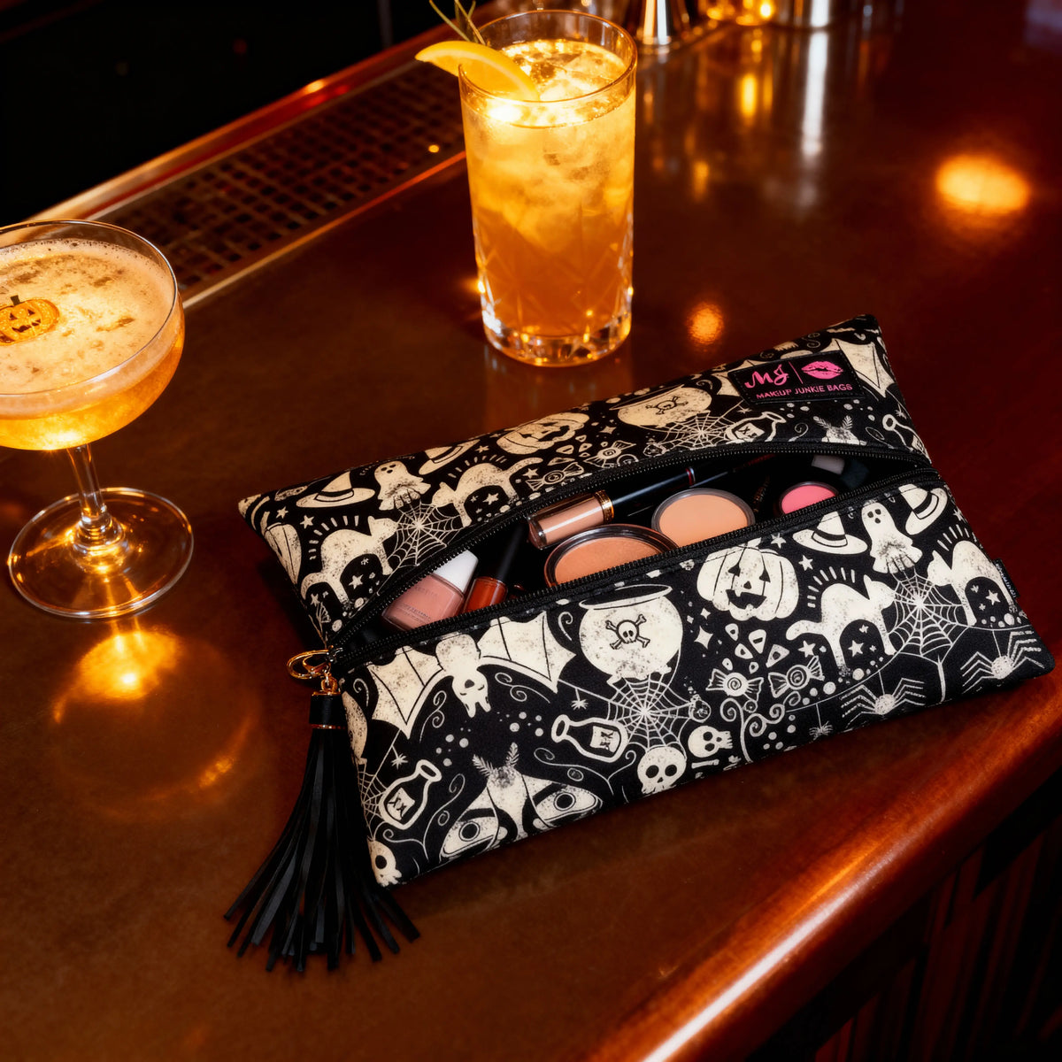 A black and white Halloween-themed makeup bag with a tassel zipper, filled with cosmetics, resting on a warm, amber-lit bar counter next to a cocktail and an iced drink.
