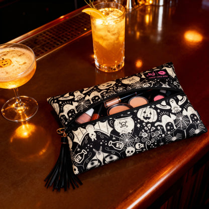 A black and white Halloween-themed makeup bag with a tassel zipper, filled with cosmetics, resting on a warm, amber-lit bar counter next to a cocktail and an iced drink.