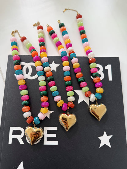 Four colorful beaded necklaces with gold heart pendants are arranged on a black card with white lettering. The necklaces feature multicolored round beads and shiny gold heart charms.