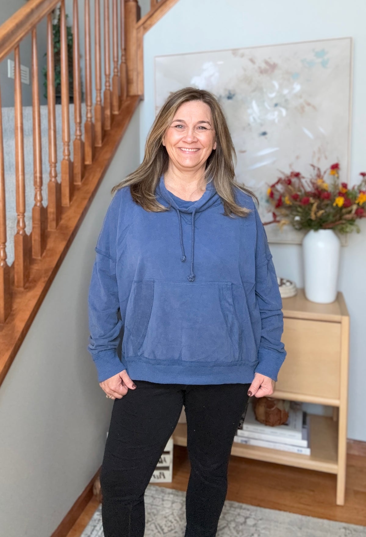 A woman wearing a dusty blue pullover hoodie and black pants stands in a home hallway next to a wooden staircase, smiling at the camera.