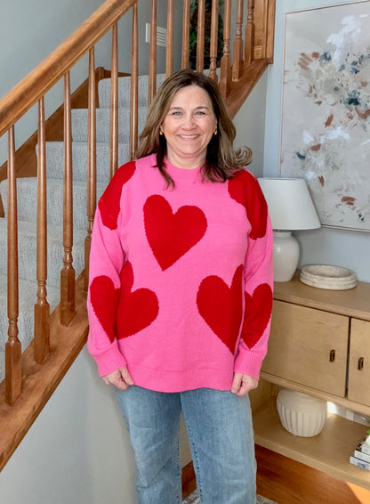 Woman wearing a pink sweater with red hearts standing in a home setting.