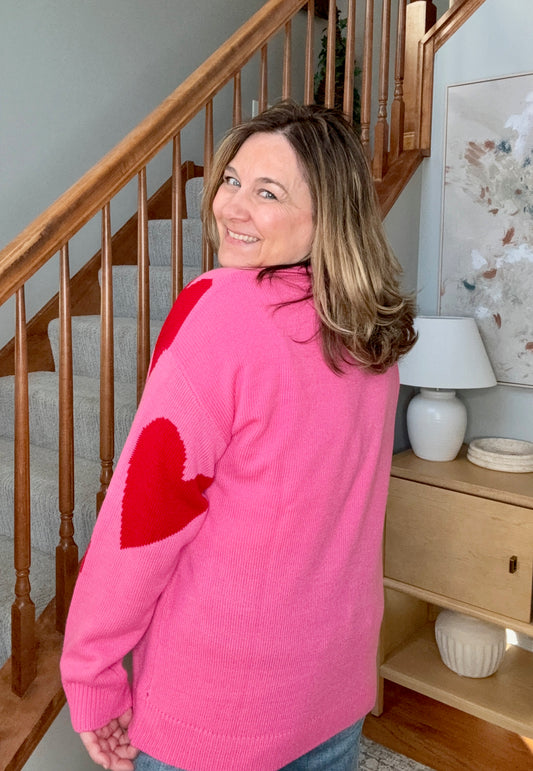 Woman wearing a pink sweater with a red heart design, standing in a home setting.