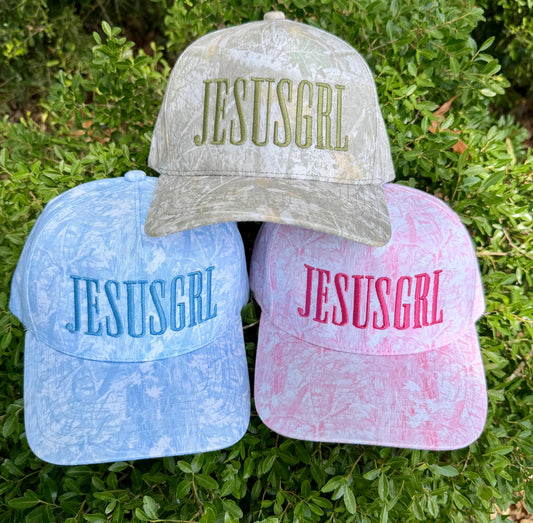 Three baseball caps with a distressed tie-dye pattern are arranged on green foliage. The top cap is light gray with olive green 'JESUSGRL' embroidery, the left cap is light blue with blue embroidery, and the right cap is pink with pink embroidery.