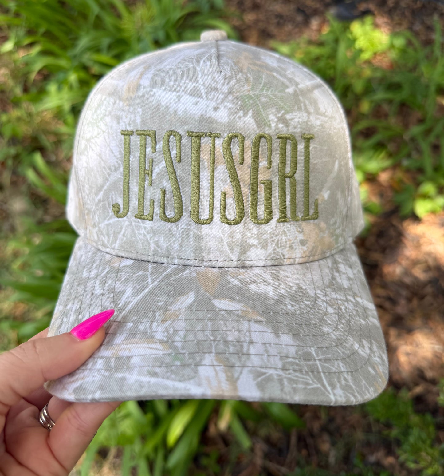 A camouflage baseball cap held by a hand with bright pink fingernails. The cap features the text 'JESUSGRL' embroidered in olive green on the front.