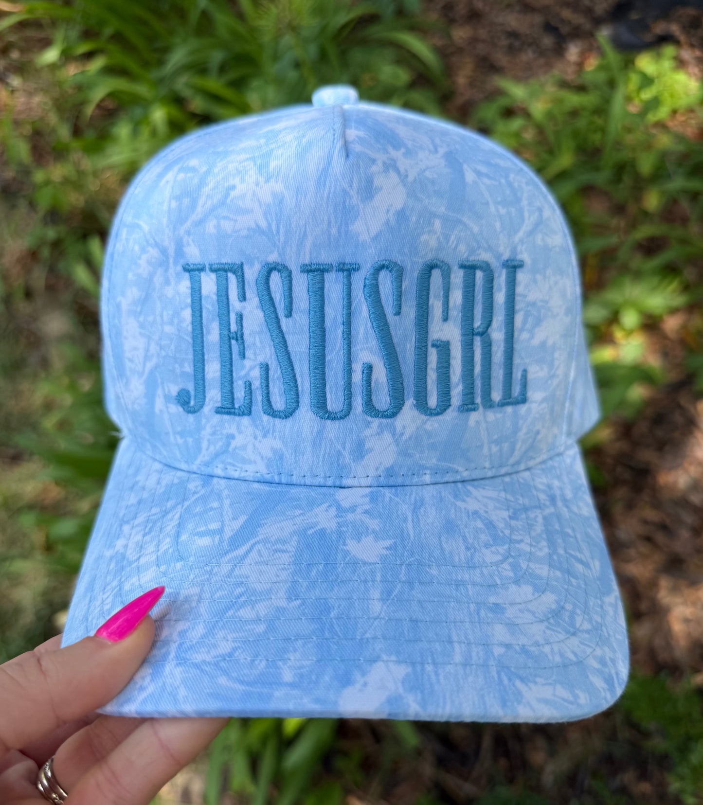A light blue marbled baseball cap with 'JESUSGRL' embroidered in blue on the front, held by a hand with pink nail polish against a blurred green outdoor background.