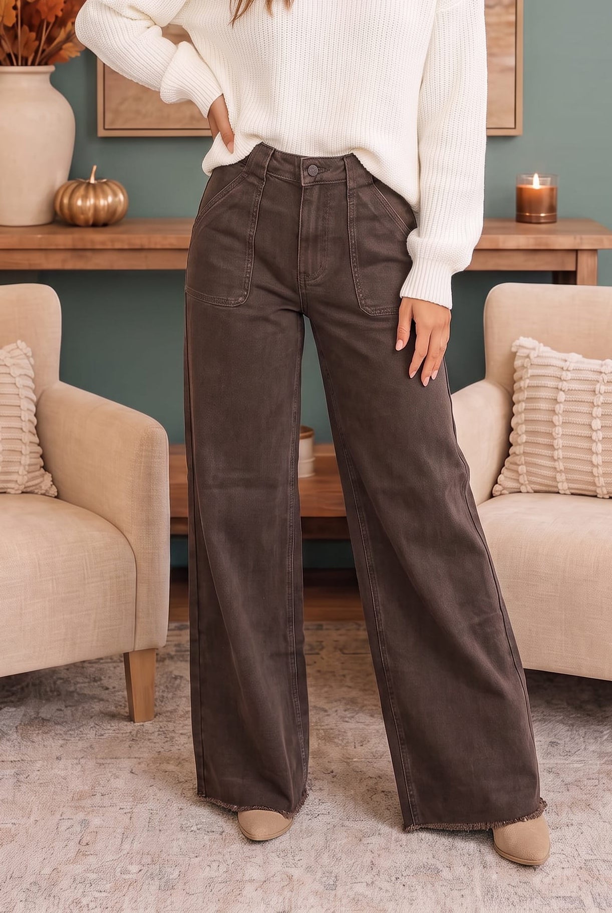 A person wearing dark brown wide-leg pants with a cream ribbed sweater, standing in a cozy living room setting with beige armchairs and a wooden console table.
