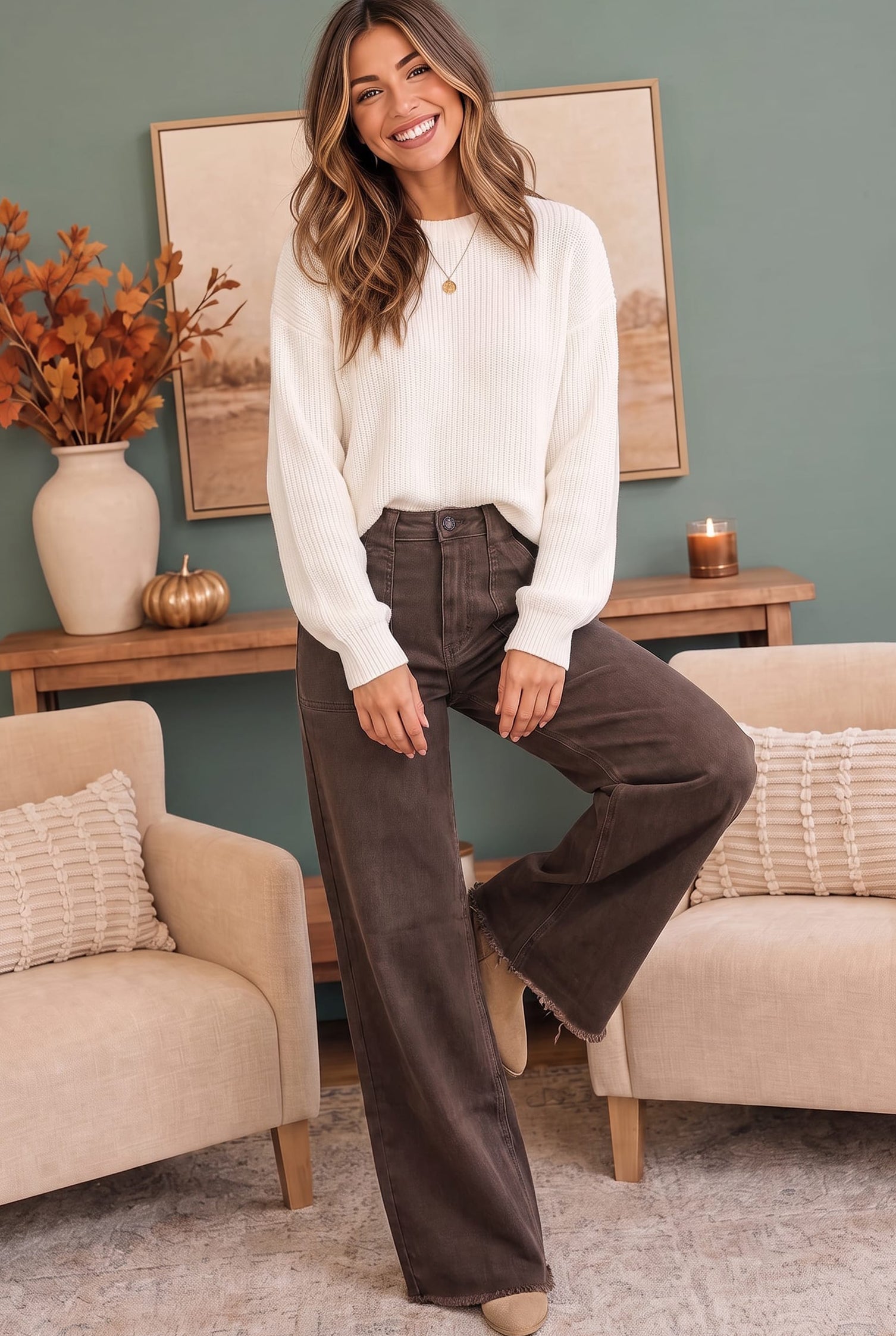 A woman stands in a cozy living room wearing a cream ribbed knit sweater and dark brown wide-leg pants. The room features a sage green wall, a wooden console table with a vase of autumn leaves, and two beige armchairs.