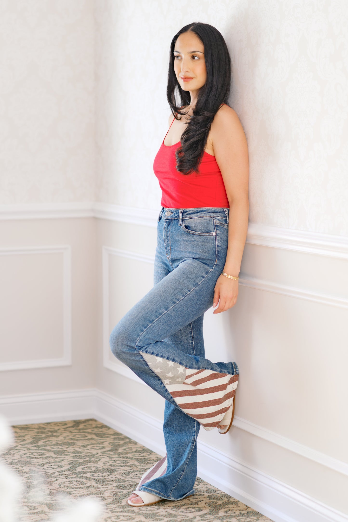 Woman wearing a red top and blue jeans leaning against a white wall.