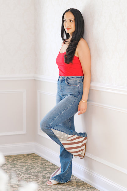 Woman wearing a red top and blue jeans leaning against a white wall.