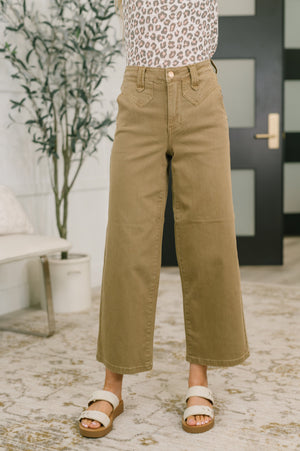 Person wearing tan wide-leg pants in an indoor setting with a plant and door in the background.