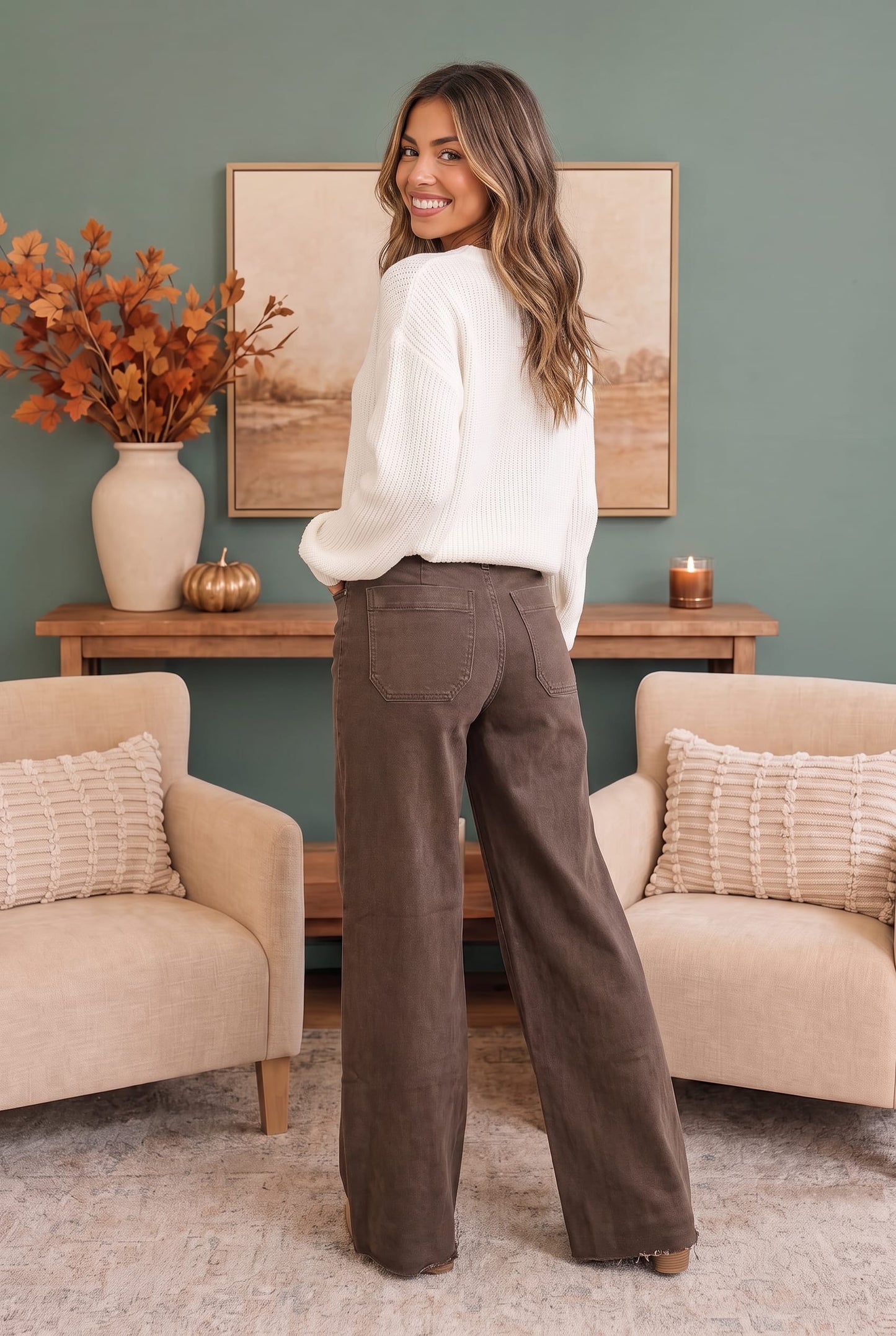 A woman stands in a living room wearing a white knit sweater and brown wide-leg pants, photographed from the back. The room has a muted green wall, a wooden console table, framed art, a vase with orange leaves, and two beige armchairs with textured pillows.