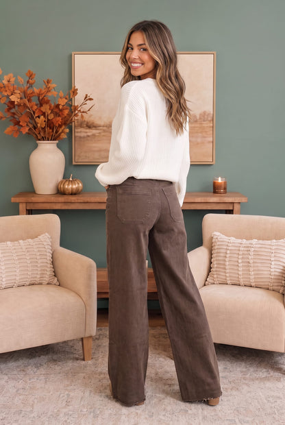 A woman stands in a living room wearing a white knit sweater and brown wide-leg pants, photographed from the back. The room has a muted green wall, a wooden console table, framed art, a vase with orange leaves, and two beige armchairs with textured pillows.
