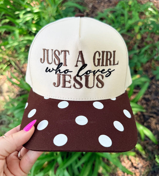A hand with pink nail polish holds a baseball cap outdoors against a blurred green foliage background. The cap has a cream-colored crown with brown and black embroidered text reading 'JUST A GIRL who loves JESUS' and a dark brown brim covered in white polka dots.