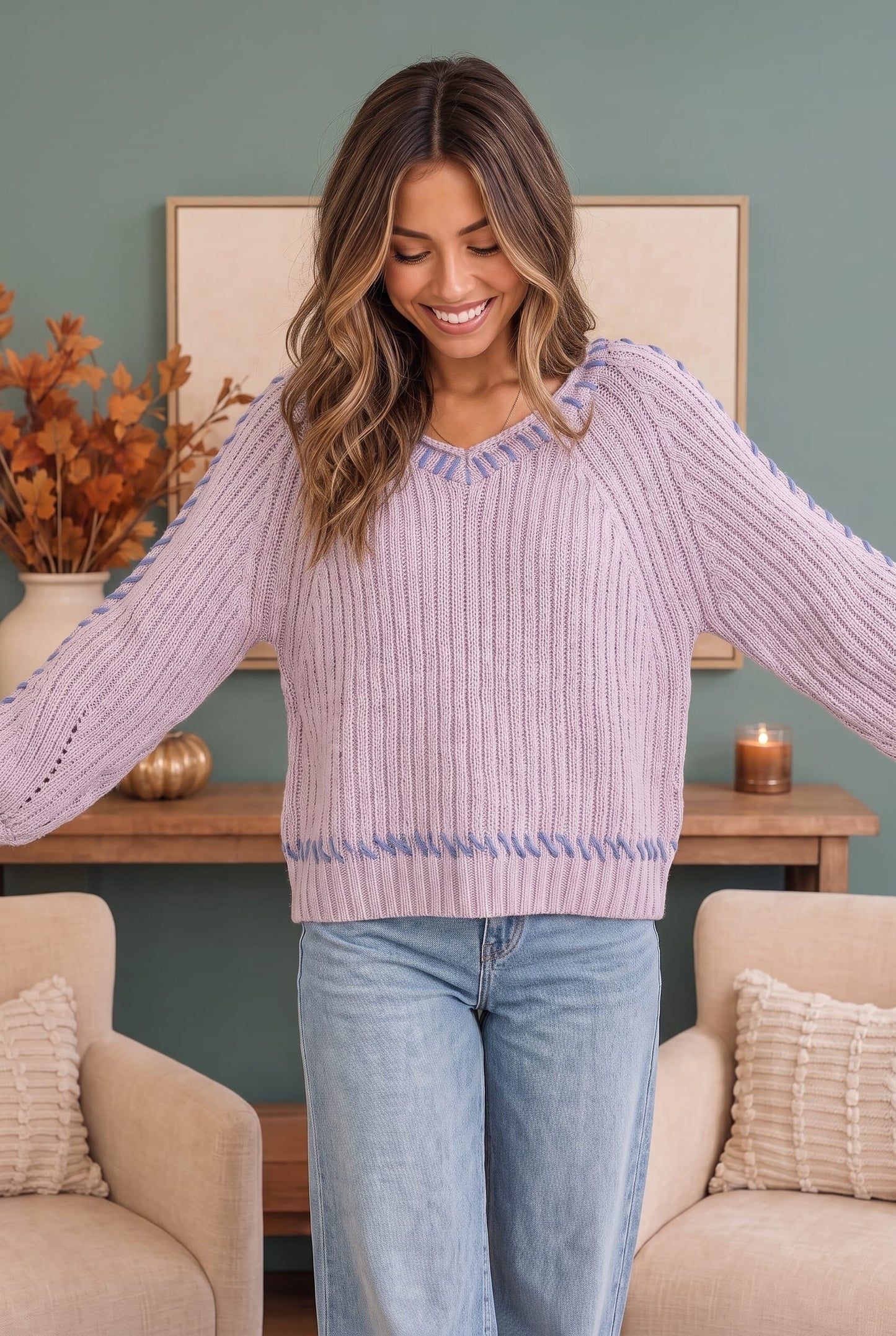 A woman wearing a lavender ribbed knit sweater with blue trim on the shoulders and hem, paired with light blue jeans, standing in a cozy living room setting.
