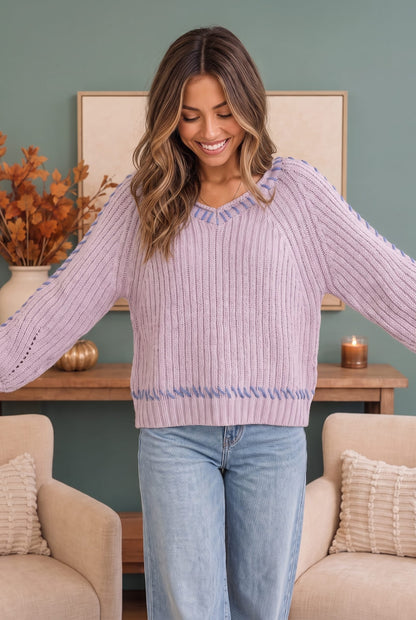 A woman wearing a lavender ribbed knit sweater with blue trim on the shoulders and hem, paired with light blue jeans, standing in a cozy living room setting.