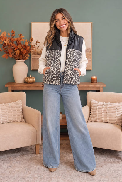 A woman stands in a living room wearing a sleeveless leopard-print vest with black faux-leather shoulder panels, a white long-sleeve shirt, and light blue wide-leg jeans.