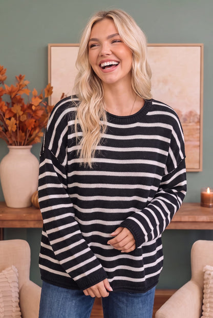 A woman with blonde hair wearing a black and white horizontally striped knit sweater and blue jeans, standing in a cozy living room setting.