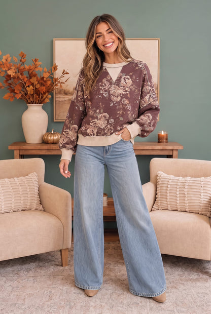 A woman stands in a styled living room wearing a brown floral long-sleeve sweatshirt with beige ribbed trim and light blue wide-leg jeans.