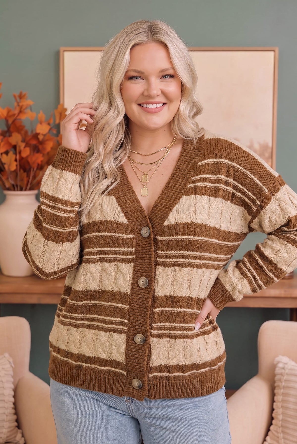 A woman wearing a brown and beige striped cable-knit cardigan sweater with button-front closure, paired with light blue jeans, standing in a cozy indoor setting.