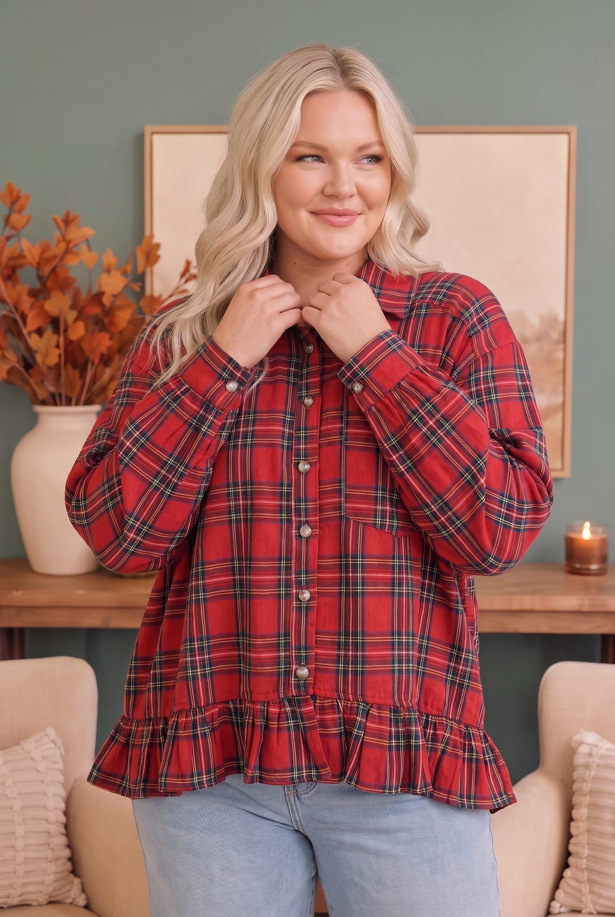 A woman wearing a red and black plaid long-sleeve button-up shirt with a ruffled hem, paired with light blue jeans, standing in a cozy living room setting.