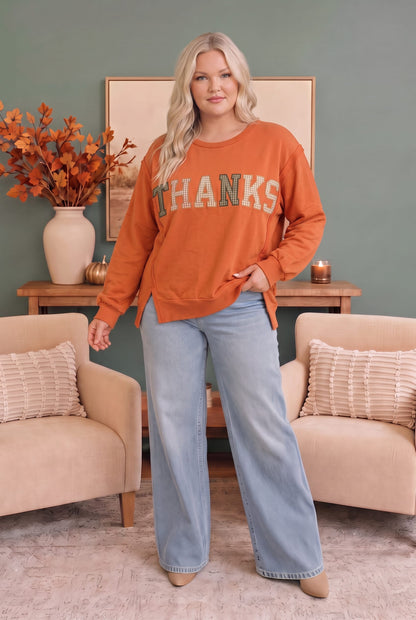 A woman stands in a styled living room wearing a burnt orange crewneck sweatshirt with 'THANKS' printed across the front in teal and white, paired with light blue wide-leg jeans.