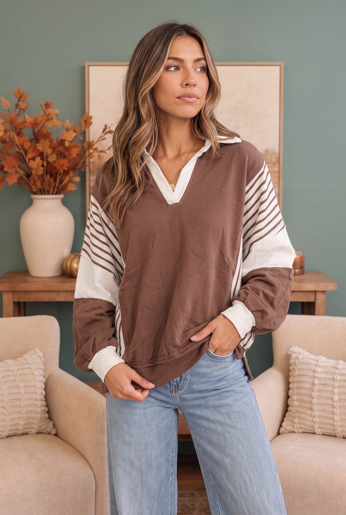 A woman wearing a brown and white color-blocked long-sleeve pullover sweatshirt with striped details on the sleeves, paired with light blue jeans, standing in a cozy living room setting.