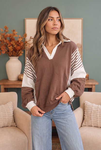 A woman wearing a brown and white color-blocked long-sleeve pullover sweatshirt with striped details on the sleeves, paired with light blue jeans, standing in a cozy living room setting.