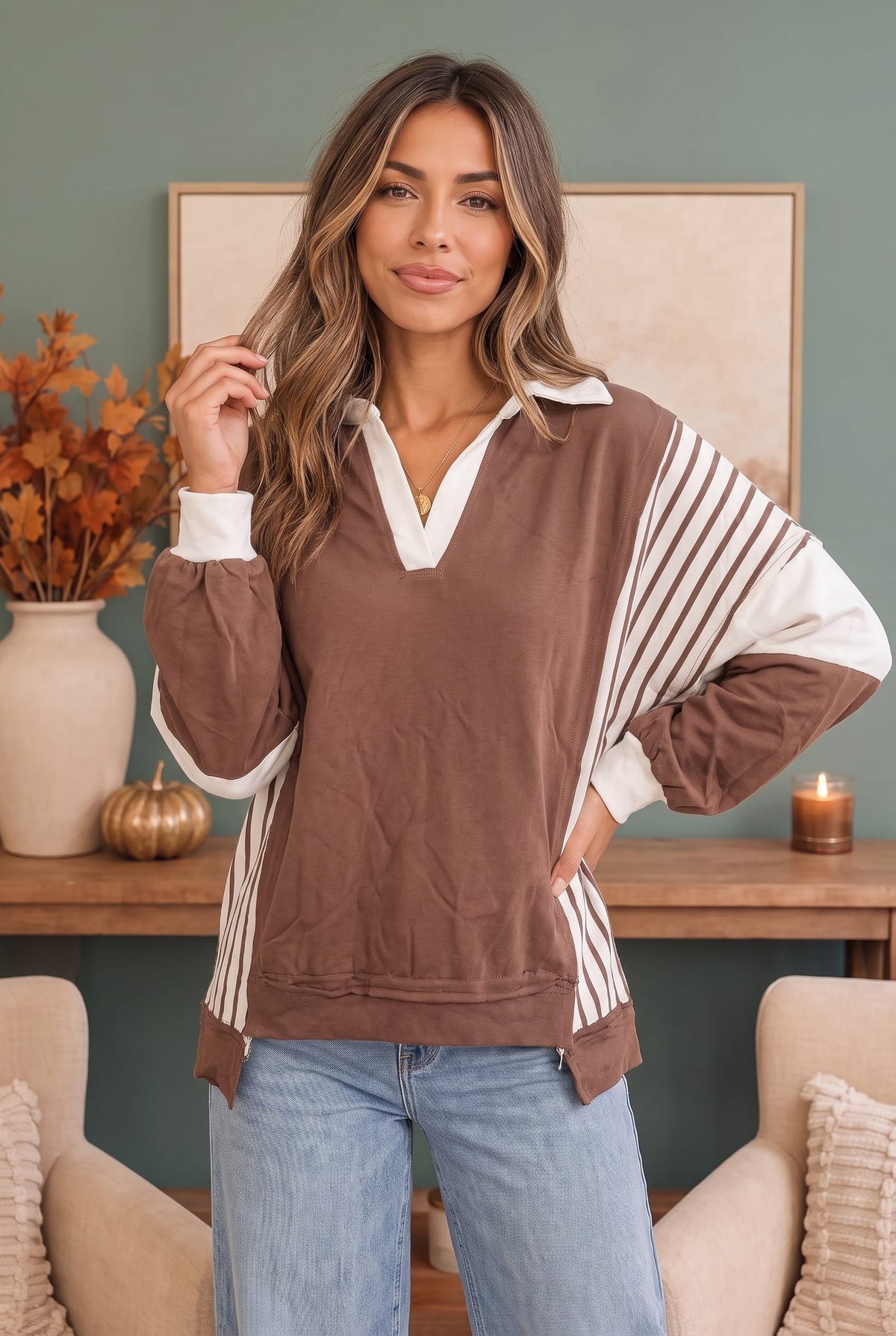 A woman modeling a brown and white color-blocked long-sleeve collared top with striped side panels, paired with light blue jeans in a cozy indoor setting.