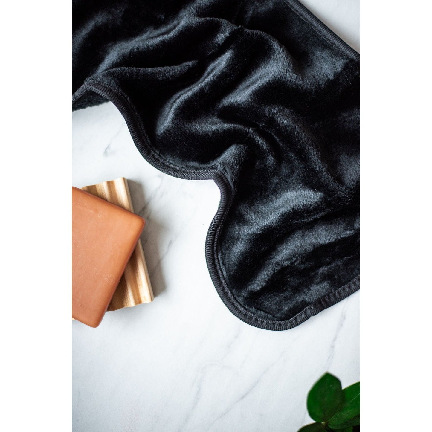 Black velvet eye mask on a white surface with a gold product and green leaves in the background