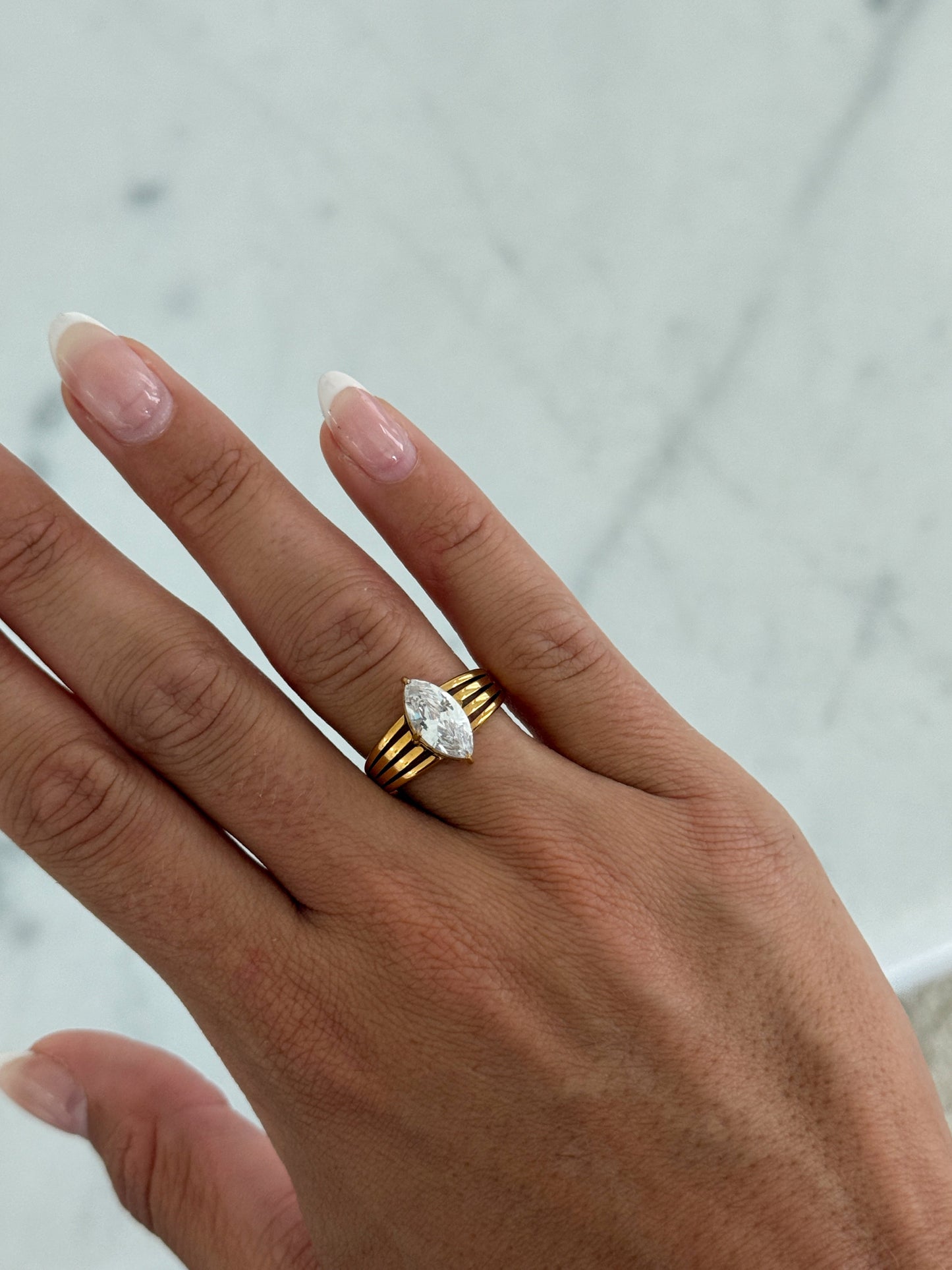 A close-up of a hand wearing a gold ring with a marquise-cut clear gemstone, set against a light marble background.