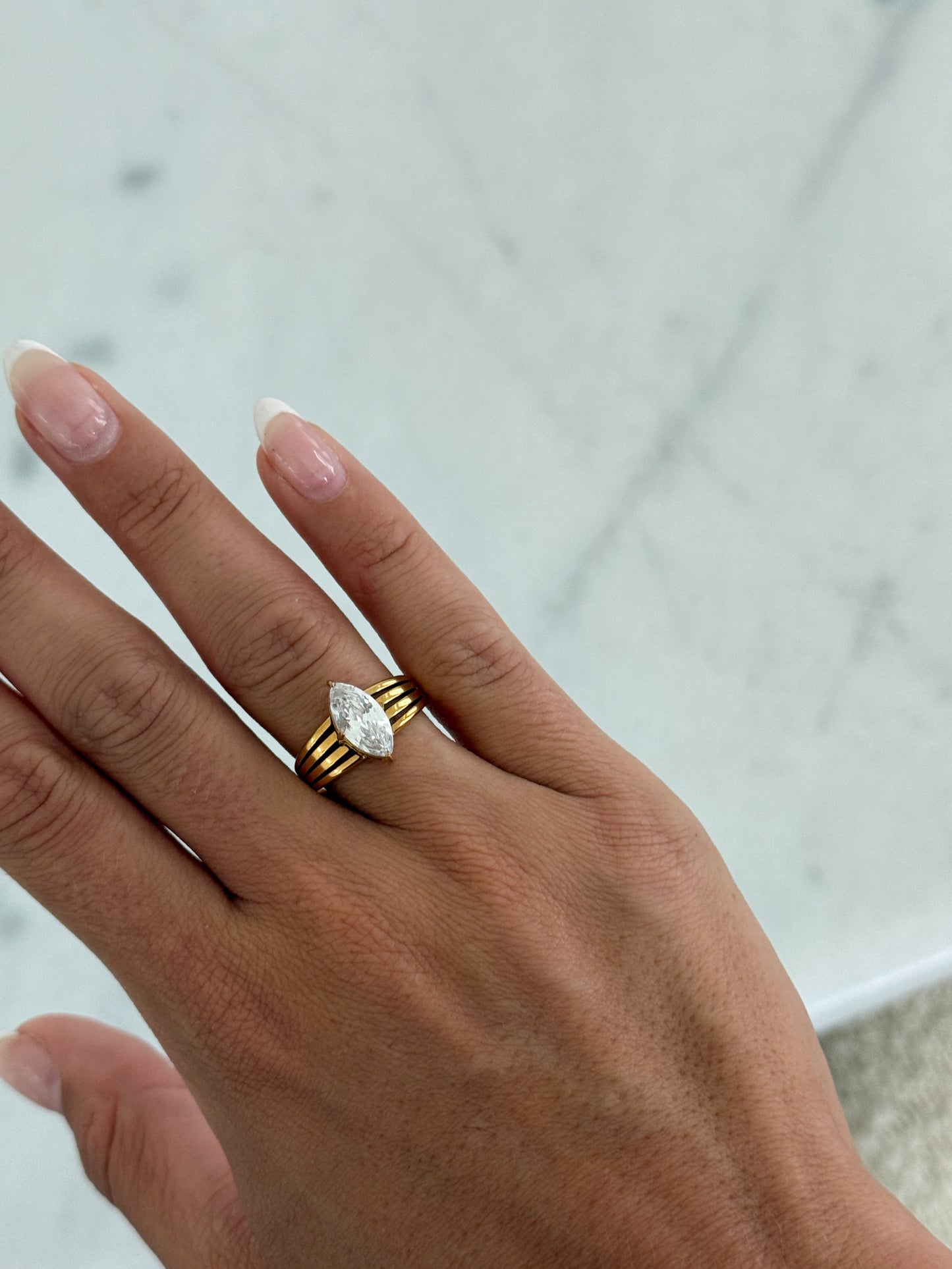 A close-up of a hand wearing a gold ring with a marquise-cut clear gemstone, set against a light marble background.