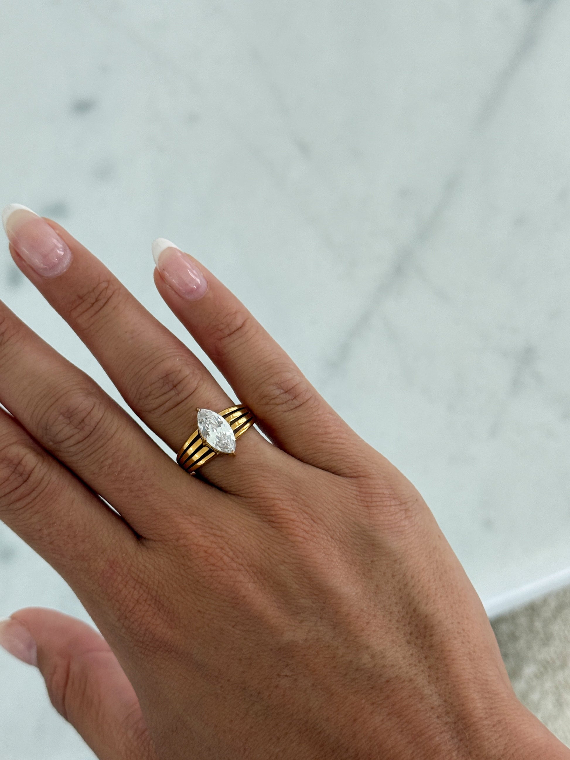 A close-up of a hand wearing a gold ring with a marquise-cut clear gemstone, set against a light marble background.