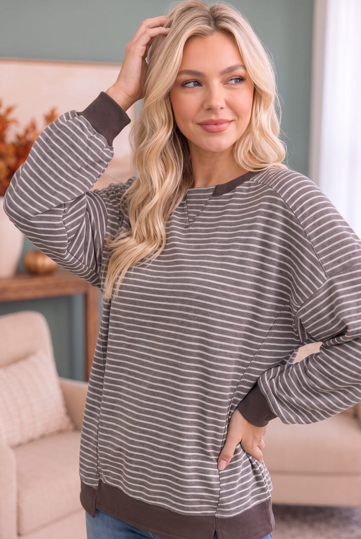 A woman modeling a long-sleeve crewneck sweatshirt with thin brown and gray horizontal stripes, featuring solid brown ribbed cuffs, hem, and collar, set in a cozy living room environment.