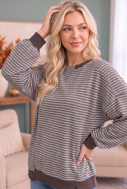 A woman modeling a long-sleeve crewneck sweatshirt with thin brown and gray horizontal stripes, featuring solid brown ribbed cuffs, hem, and collar, set in a cozy living room environment.