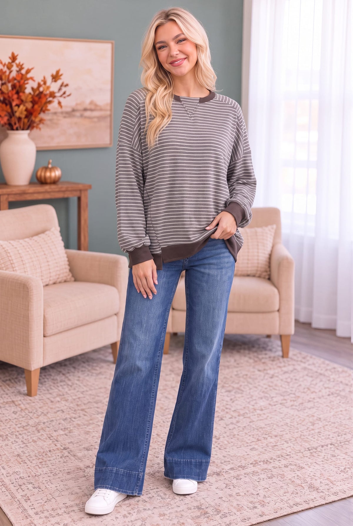 A woman stands in a living room wearing a gray and white striped long-sleeve top with dark brown ribbed trim, blue flare jeans, and white sneakers.