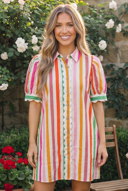 Woman wearing a colorful striped dress standing outdoors with flowers and a stone wall in the background