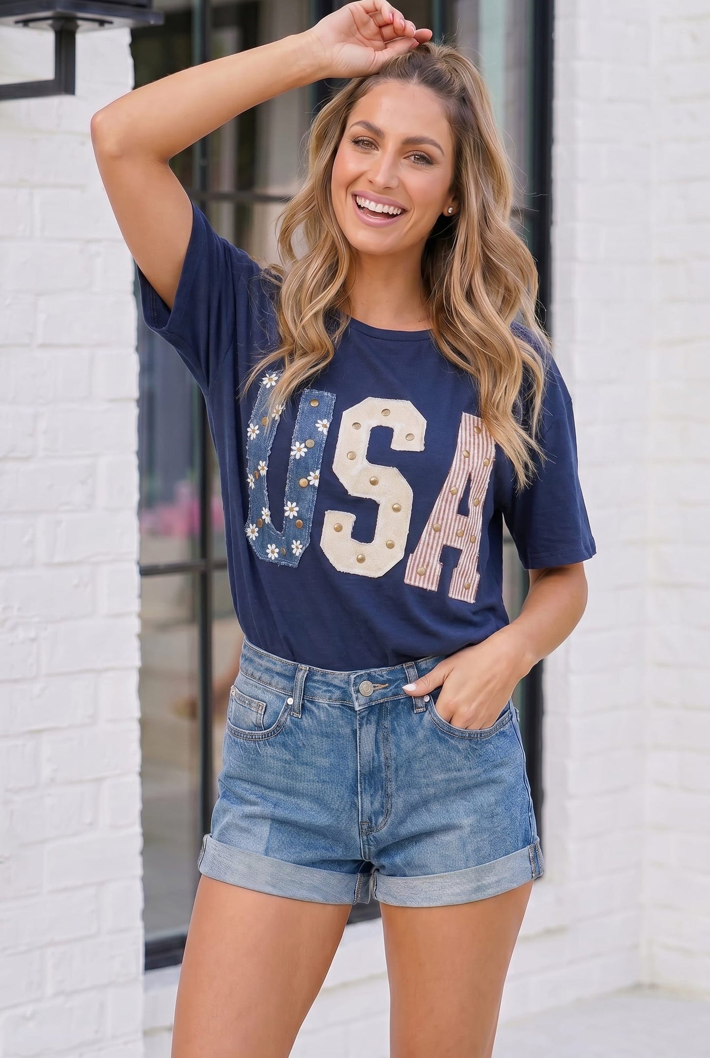 Woman wearing a navy blue 'USA' t-shirt with stars and denim shorts standing against a white brick wall.
