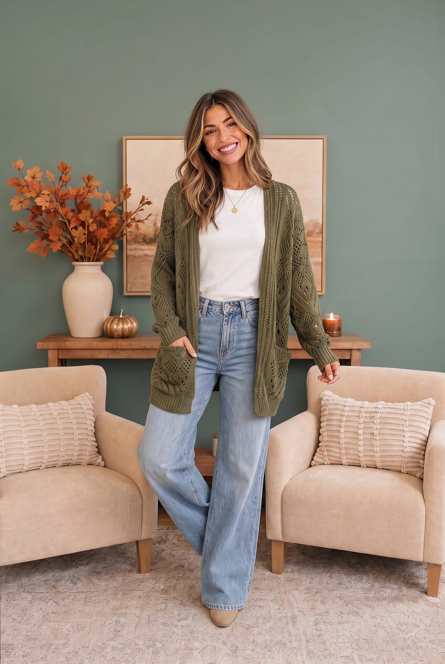 A woman stands in a styled living room wearing an olive green cable-knit cardigan over a white top and light-wash wide-leg jeans. The room features a sage green wall, a wooden console table with a vase of autumn leaves, and two beige armchairs.