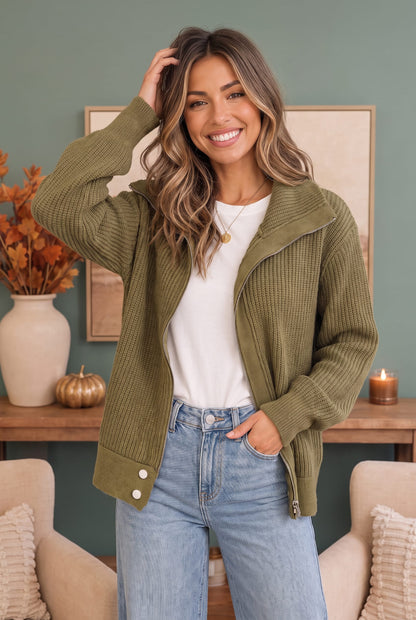 A woman wearing an olive green ribbed knit zip-up jacket over a white t-shirt and light blue jeans, standing in a cozy room with a green wall, wooden console table, and neutral chairs.