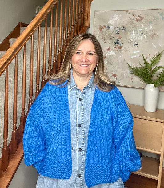 Woman wearing a blue knitted cardigan standing in a home setting with a staircase and artwork in the background.