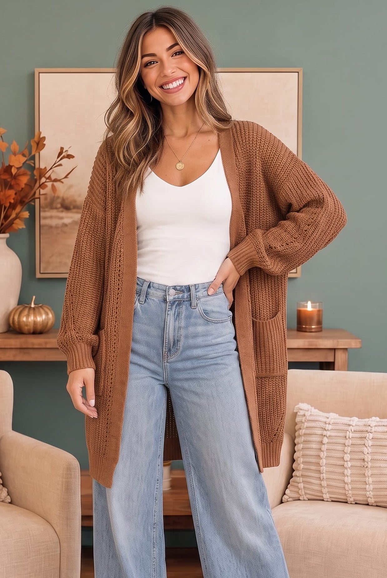 A woman stands in a living room wearing a brown open-front ribbed knit cardigan over a white tank top and light blue wide-leg jeans.