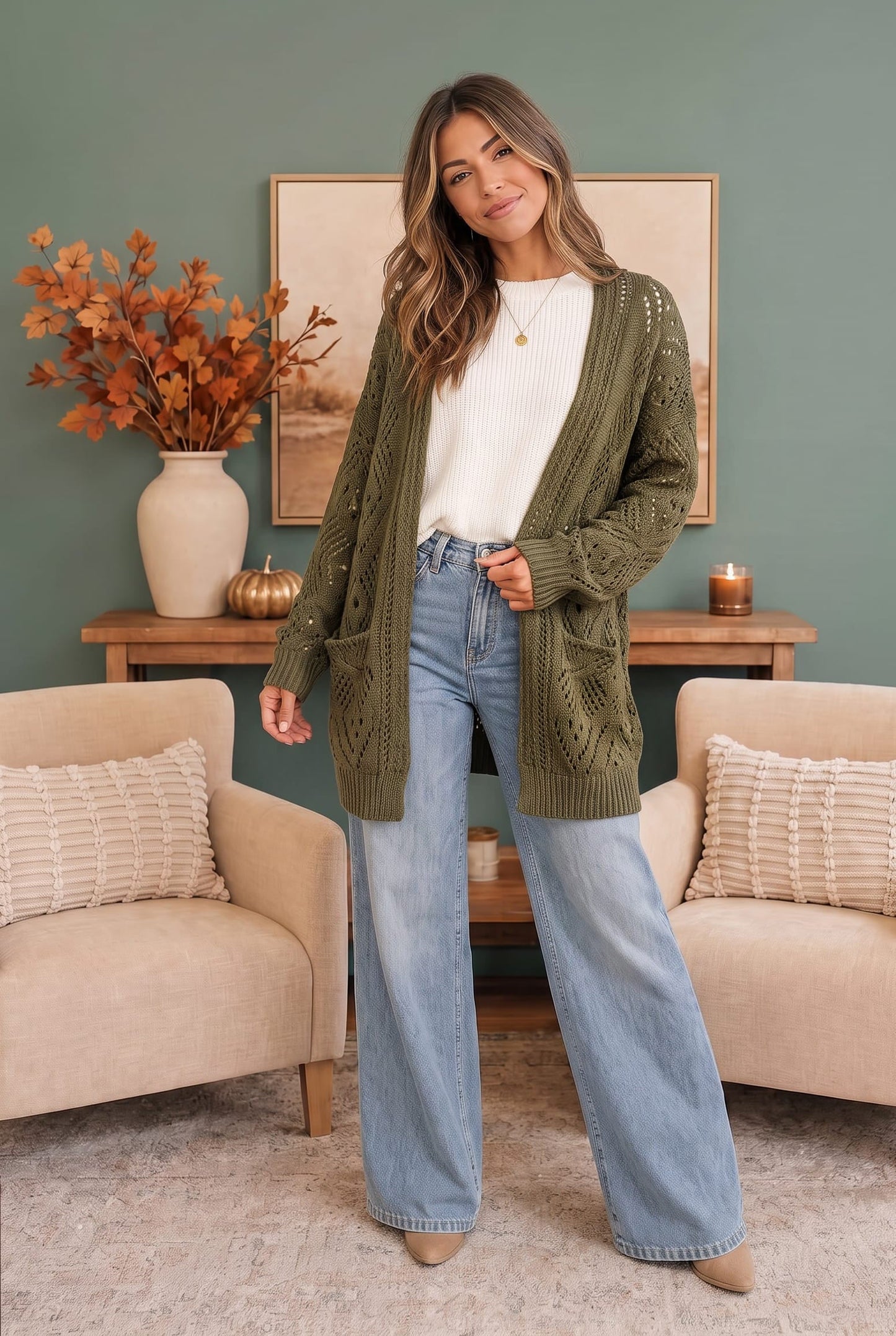 A woman stands in a styled living room wearing an olive green cable-knit cardigan over a white top and light blue wide-leg jeans.