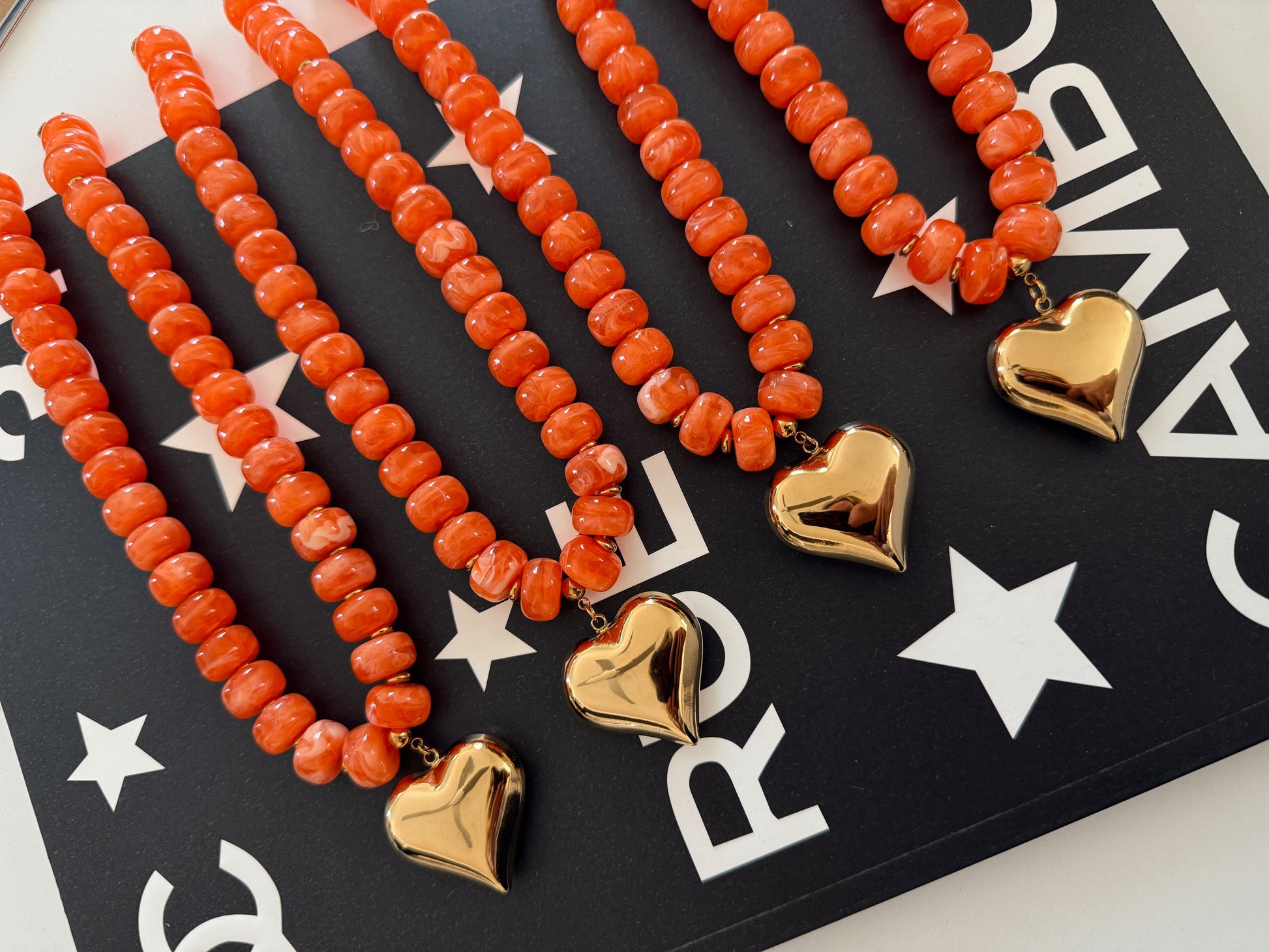 Four coral-orange beaded necklaces with gold heart charms, arranged diagonally on a black card with white stars and letters.
